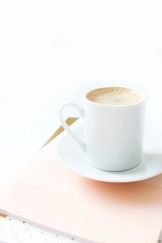 White coffee cup on a saucer atop a pale pink notebook