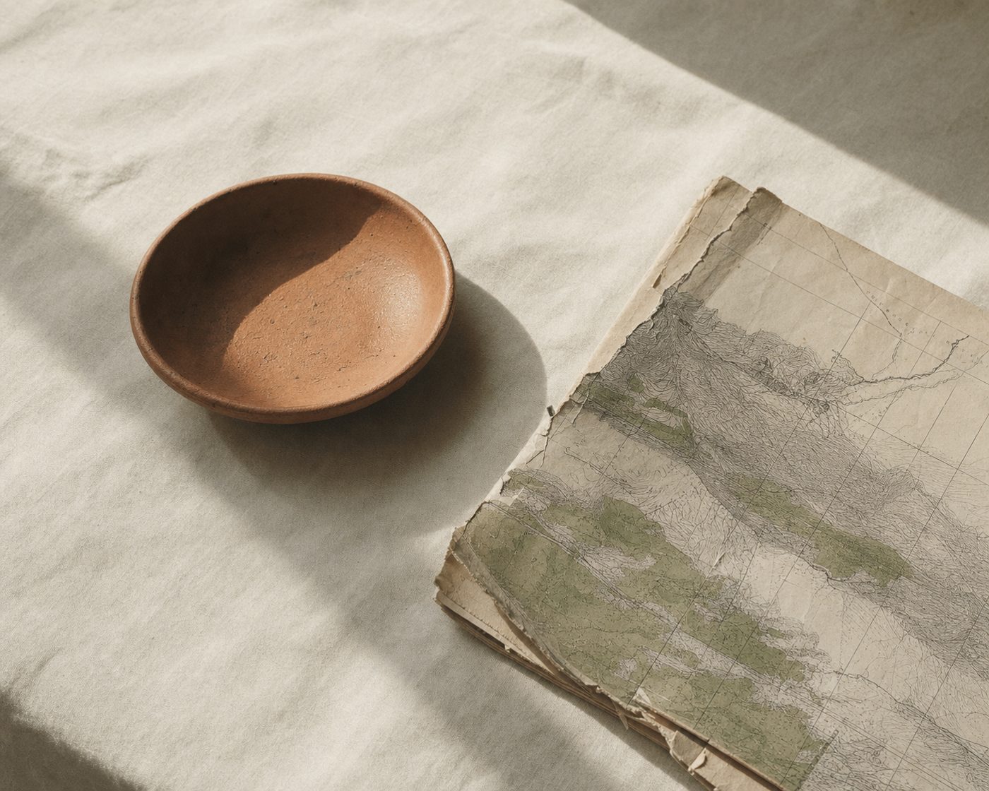 A torn folded paper map and a terracotta clay dish on cream linen in morning light